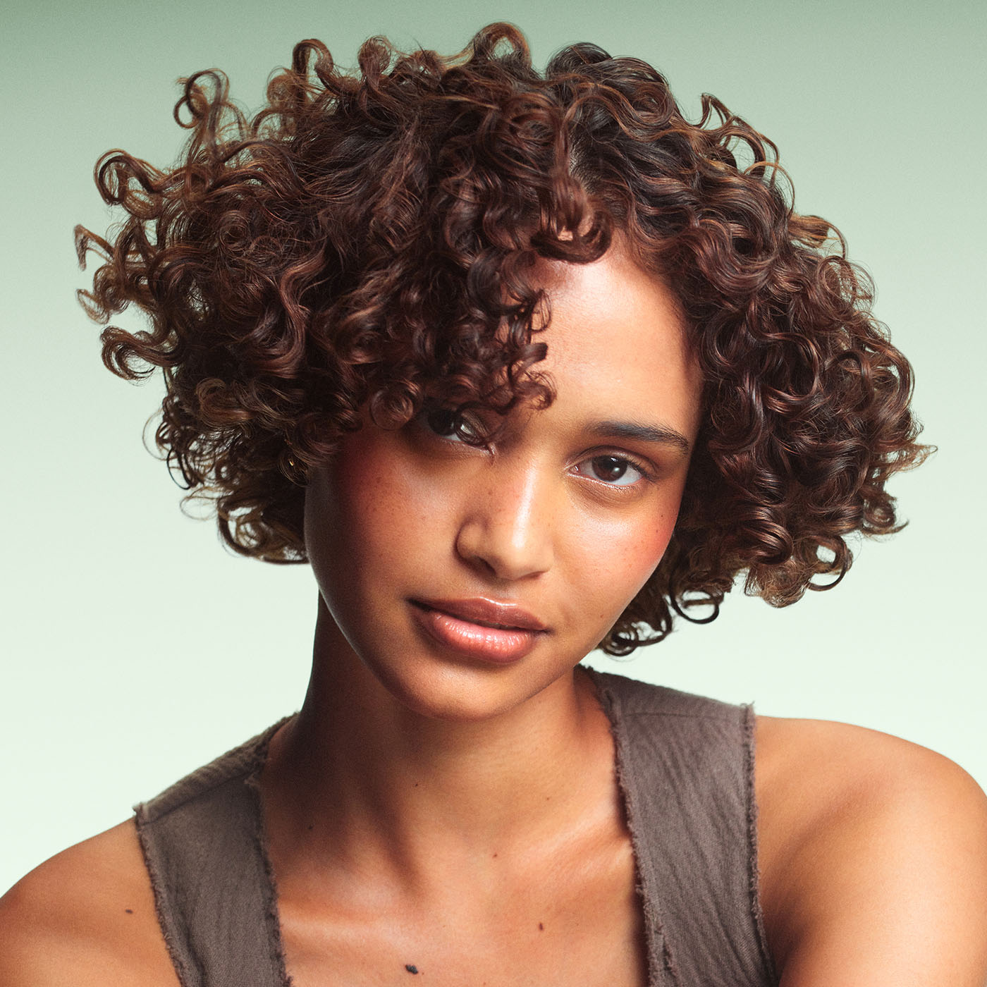 Model with short, curly brown glossy hair on green background.